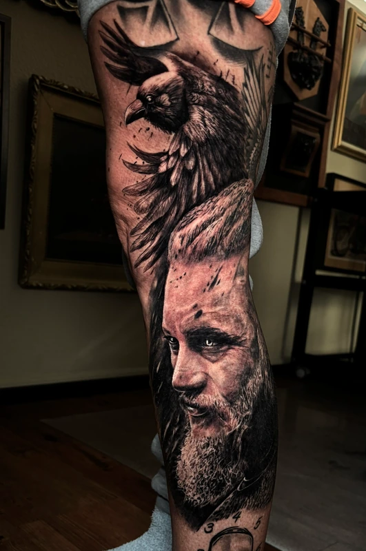 Ragnar e corvo tatuados na perna em preto e cinza